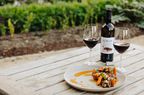 Two glasses of red wine, plate of food and Roblar wine bottle on a table outdoors.