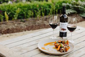 Two glasses of red wine, plate of food and Roblar wine bottle on a table outdoors.