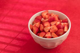 Bowl of chopped strawberries on a red surface with shadows.